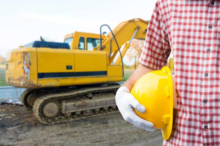 midsection-male-worker-holding-yellow-hardhat-while-standing-against-earth-mover-construction-site